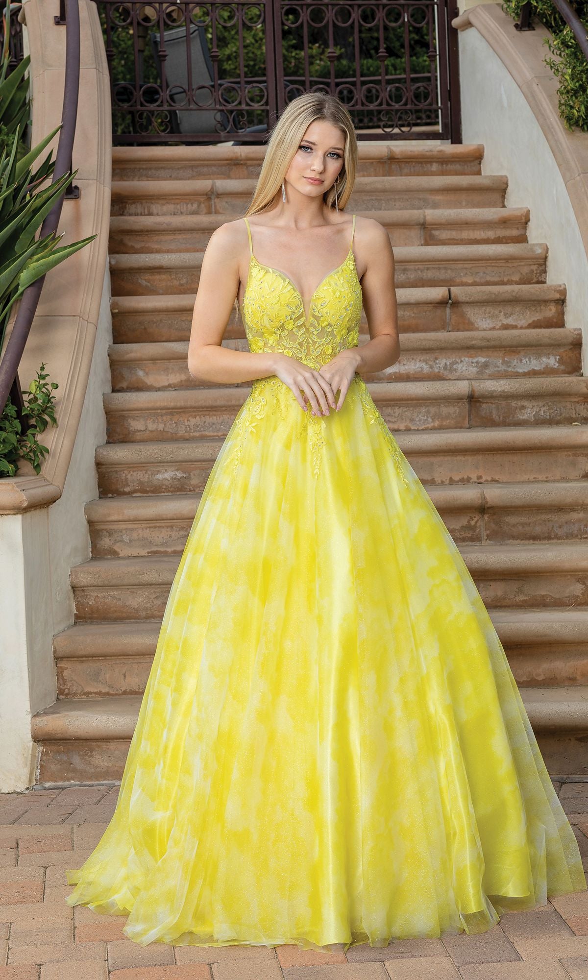 A woman in an affordable Tie-Dye Prom Ball Gown with sheer sides—a long, sleeveless dress with a fitted bodice and flowing skirt—stands on stone steps with wrought iron railings.